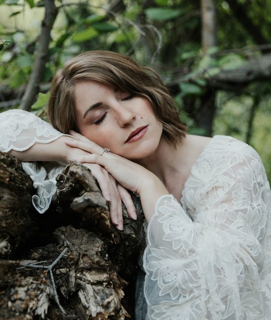 Close-up of a woman in a white lace robe resting her cheek on her hands against a mossy log in a Utah forest with eyes closed in peaceful surrender during a women's wellness retreat